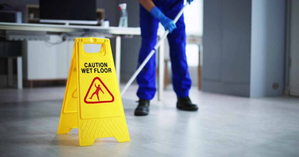 A person mopping the floor in an indoor setting provides a comprehensive solution for your health & safety needs. In the foreground, there is a yellow caution sign that reads "Caution Wet Floor," with a pictogram of a person slipping. The person mopping is dressed in blue overalls and gloves.