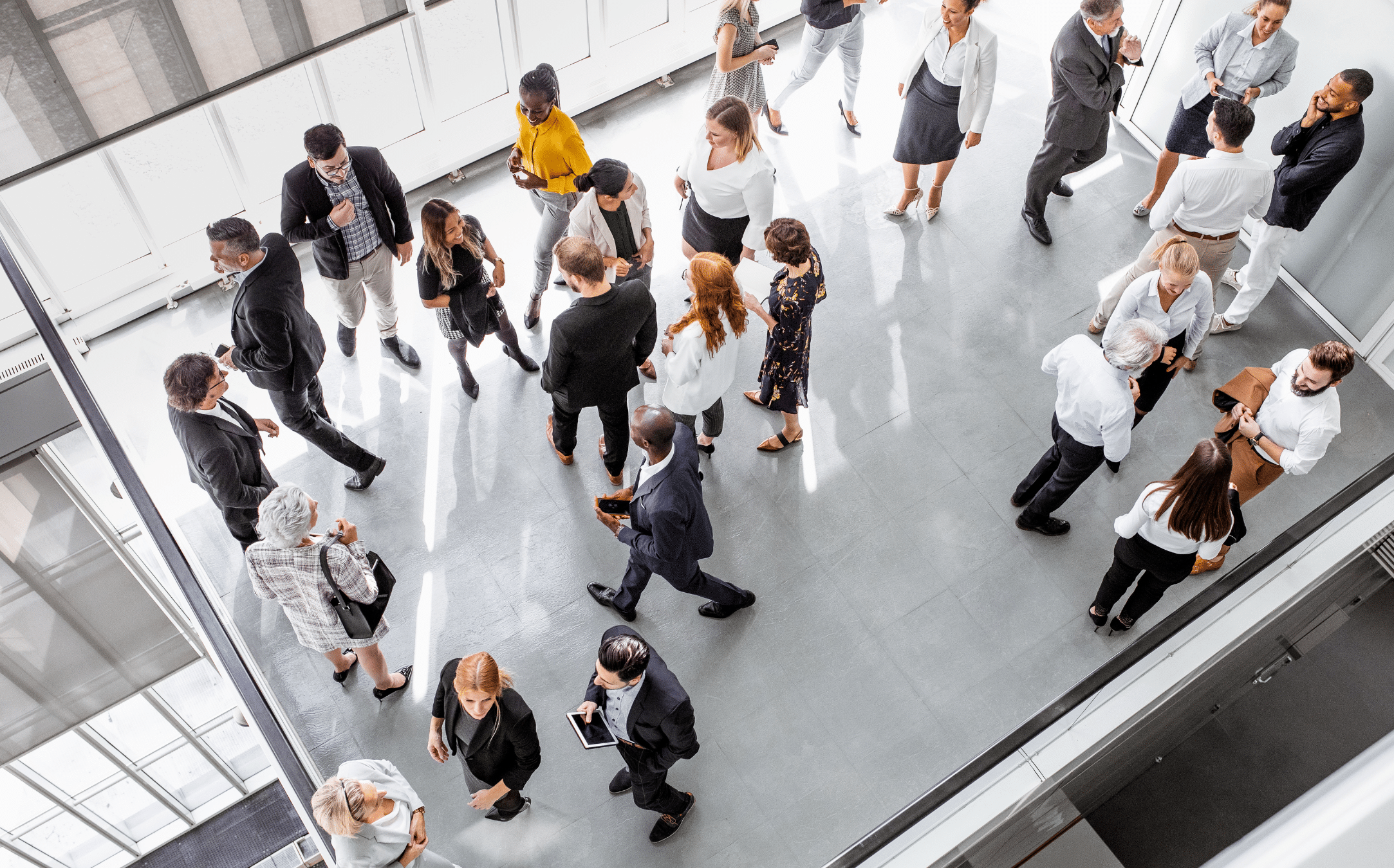 An overhead view of a modern office space bustling with activity during the ebase Spring Tradeshow Season 2024. Many people are mingling—some talking in groups, while others walk through the area. The gray floor and natural light from large windows accentuate the diverse mix of individuals.