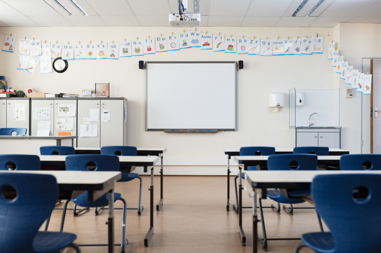 A classroom with a whiteboard at the front, surrounded by alphabet posters along the walls. Blue chairs and white desks are arranged in rows. Cabinets and a clock are on the left side, with a sink and sanitizer dispenser on the right wall.