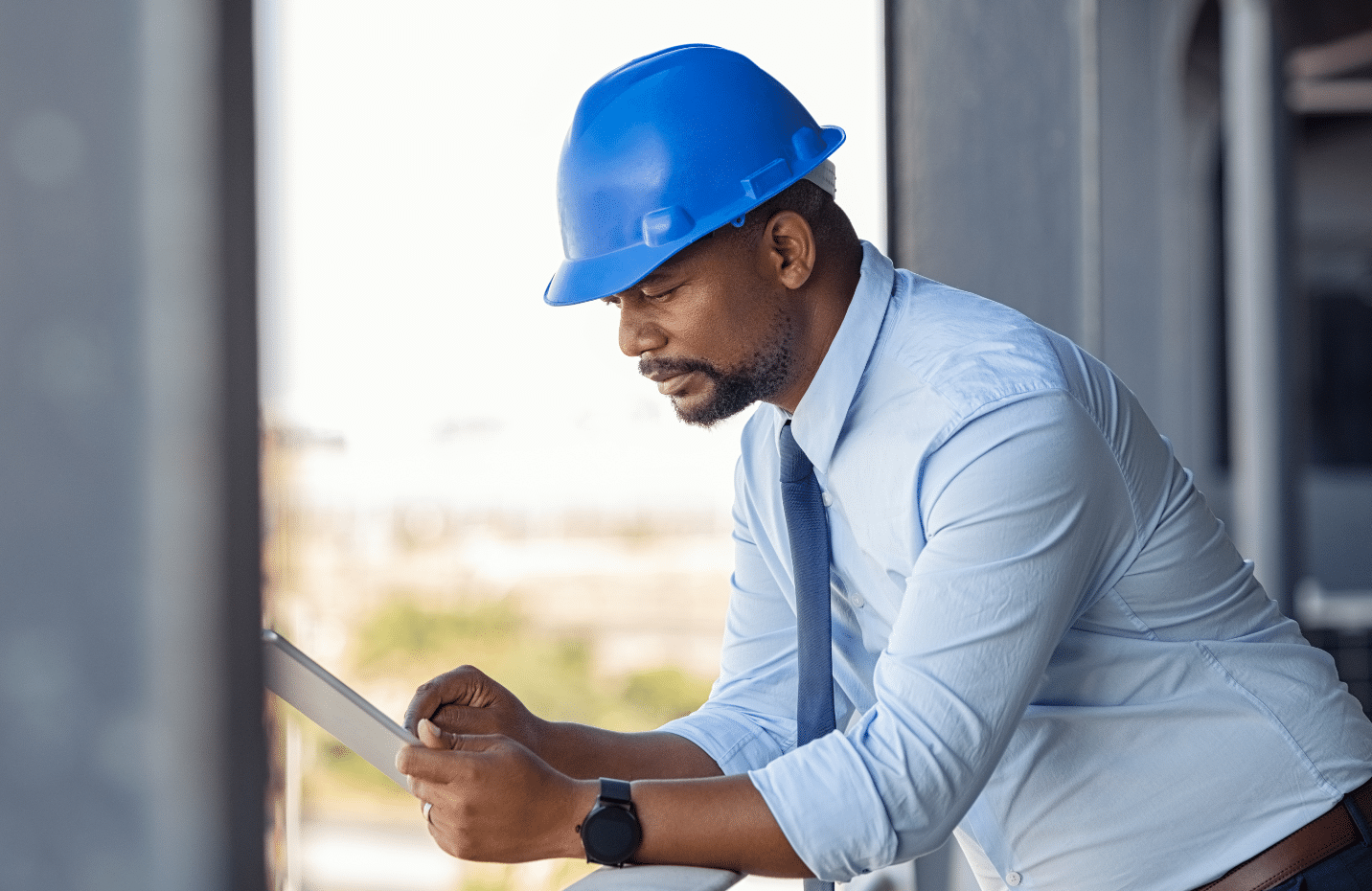 Wearing a blue hard hat and tie, the man leans on a railing, absorbed in his tablet. He appears to be analyzing an Ebase work order or construction plans. The outdoor industrial backdrop underscores his focus and expertise in managing complex data on site.