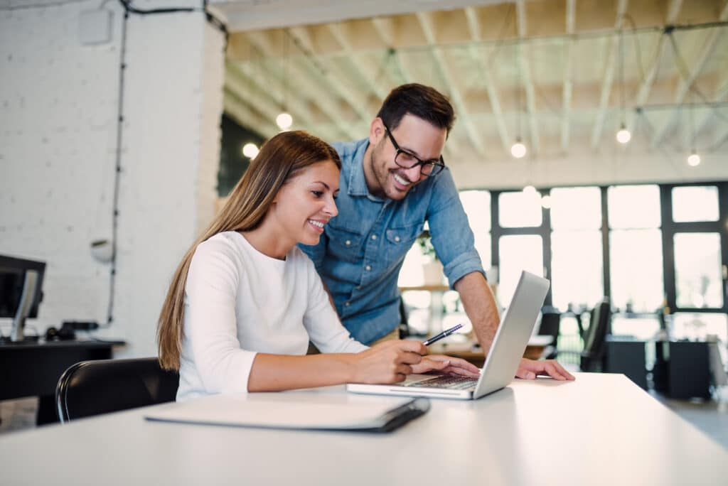 A woman and a man are smiling while working together at a desk with a laptop in a modern office. The woman is typing, and the man stands next to her holding a pen, both appearing engaged and collaborative, perhaps brainstorming ideas for smarter scheduling in the New Year.