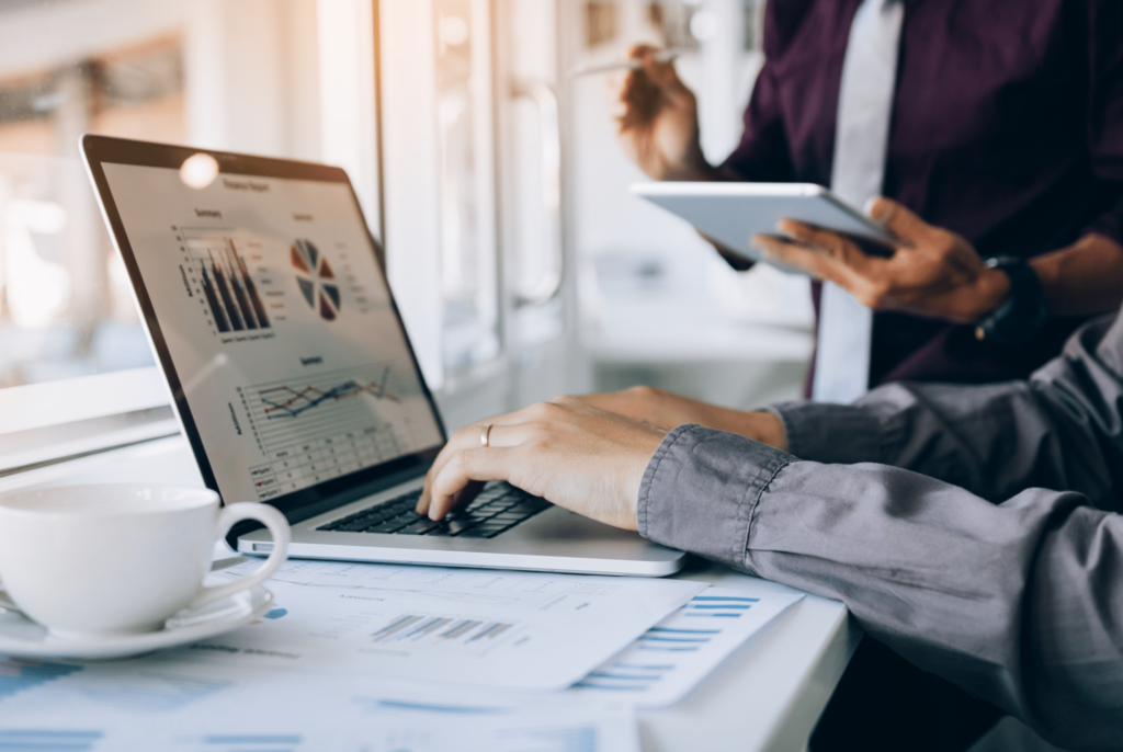 A person is working on a laptop displaying graphs and charts, hinting at future trends. Nearby, someone holds a tablet, using digital logbooks to organize data. A cup of coffee and documents with charts are on the table, suggesting a business or data analysis setting.