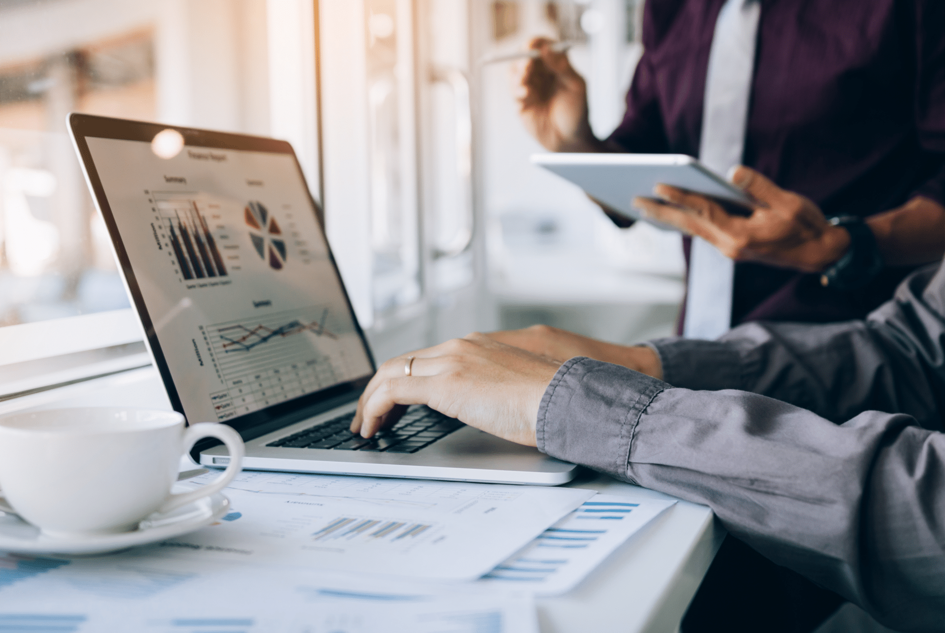 A person is working on a laptop displaying graphs and charts, hinting at future trends. Nearby, someone holds a tablet, using digital logbooks to organize data. A cup of coffee and documents with charts are on the table, suggesting a business or data analysis setting.