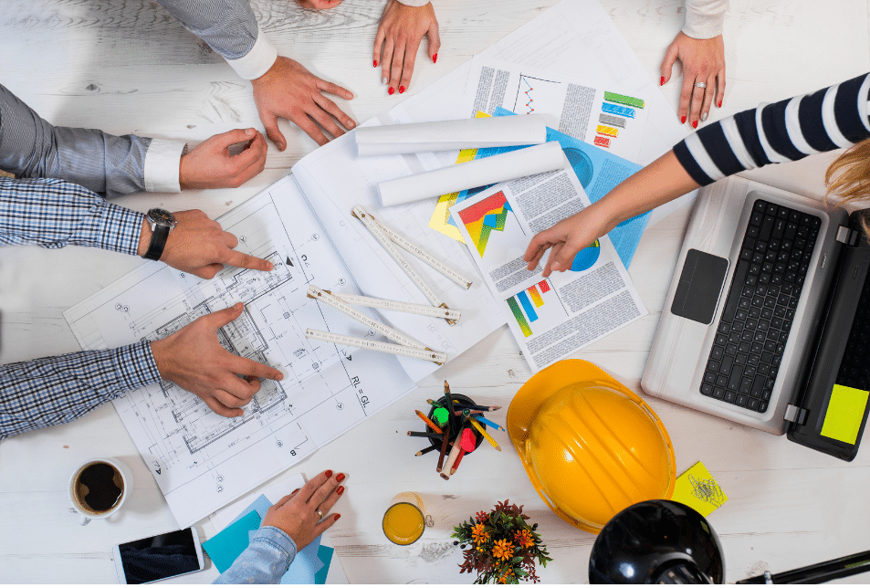 A group of people collaborating over a table filled with architectural blueprints, a laptop, vibrant charts, and a yellow hard hat. With coffee cups in hand and plans unfolding before them, hands are actively pointing and discussing how to ebase projects. A small plant adds a touch of green to the action-packed scene.