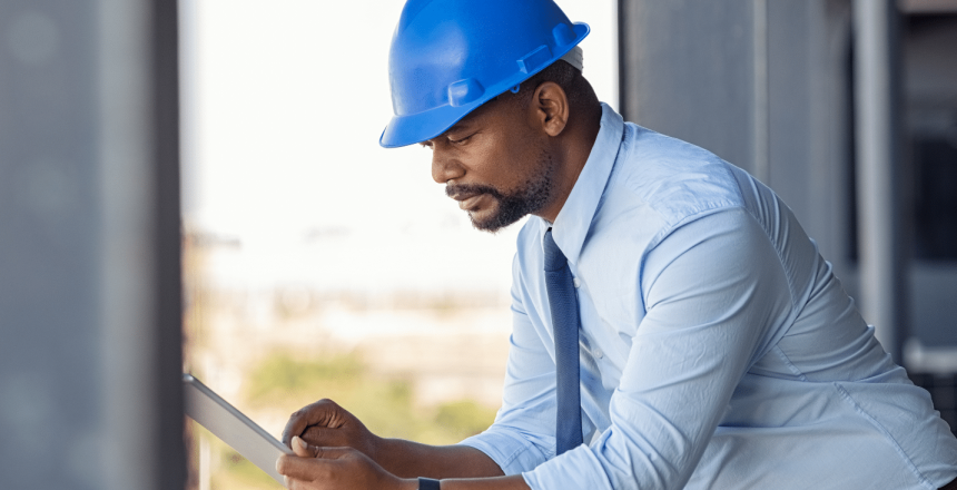 Wearing a blue hard hat and tie, the man leans on a railing, absorbed in his tablet. He appears to be analyzing an Ebase work order or construction plans. The outdoor industrial backdrop underscores his focus and expertise in managing complex data on site.