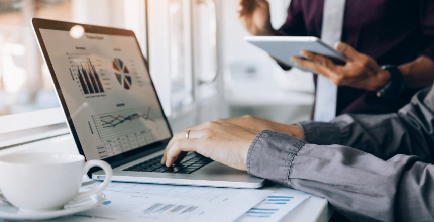 A person is working on a laptop displaying graphs and charts, hinting at future trends. Nearby, someone holds a tablet, using digital logbooks to organize data. A cup of coffee and documents with charts are on the table, suggesting a business or data analysis setting.